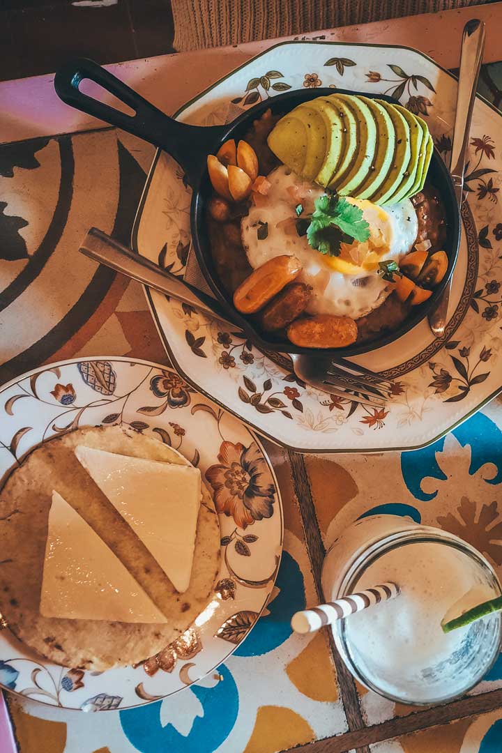 cafe-manacas-jardin Close-up of a colorful Colombian breakfast at a café in Jardín, featuring a skillet of eggs, avocado, mini arepas, and tomatoes, with a side plate of arepas and cheese and a creamy drink with a striped straw.