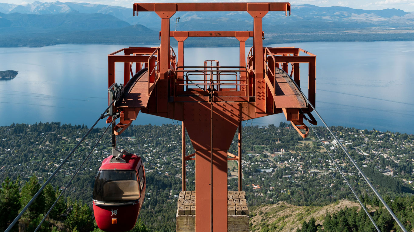 cerro-otto-bariloche A red gondola from Cerro Otto ascends over lush forest with panoramic views of Bariloche and Lake Nahuel Huapi below, surrounded by distant mountains.