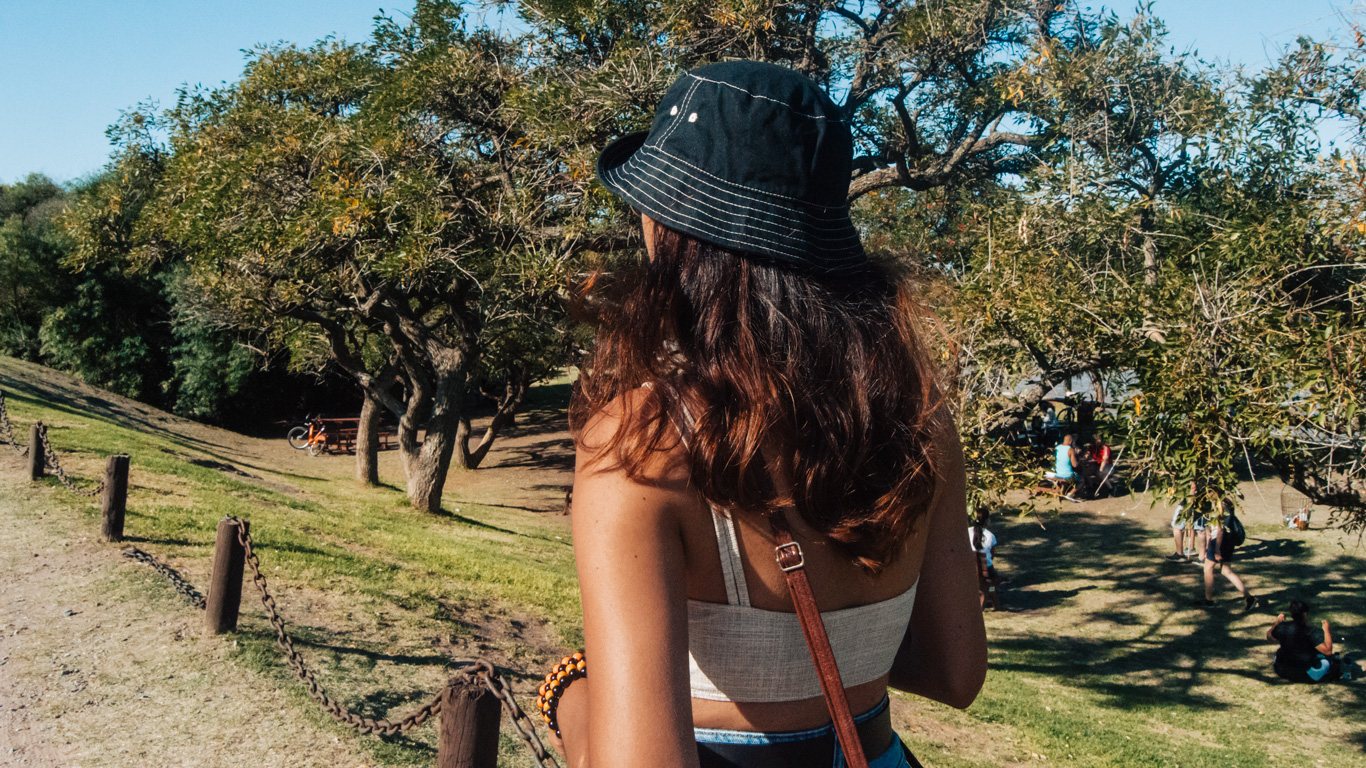costanera-sur-ecological-reserve A woman wearing a black bucket hat walks toward a grassy area shaded by trees, with small groups of people sitting or walking in the distance.