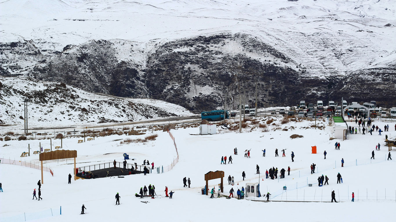 farellones-chile Tourists enjoy winter activities in Farellones, Chile during the snow season in Santiago, with snowy hills, parked vehicles, and a scenic mountain backdrop.