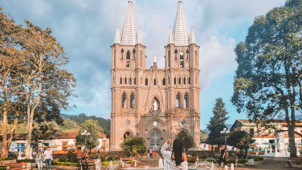 jardin-antioquia-1 A woman with long dark hair stands in a vibrant town square, facing a grand brick church with twin spires in Jardín, Antioquia, Colombia. The square is framed by tall trees, colonial-style buildings, and people walking or sitting on benches, capturing a peaceful, sunny day in a picturesque mountain town.