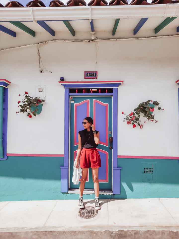 jardin-colombia-1 A stylish woman poses in front of a brightly painted colonial-style door in Jardín, Colombia, featuring vivid shades of blue, purple, and pink with flowerpots flanking the entrance.