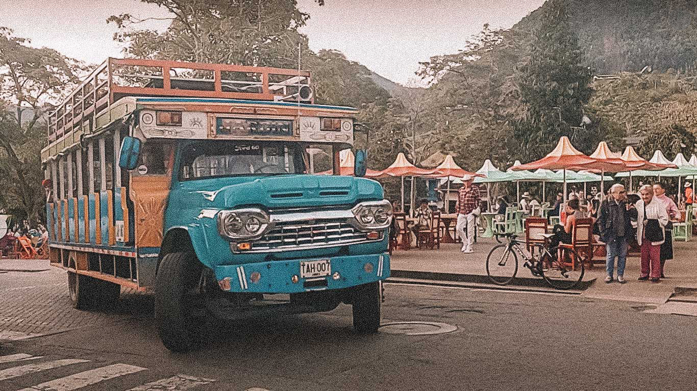 jardin-colombia-chiva A brightly painted traditional Colombian “chiva” bus in shades of blue, orange, and white drives through a lively street in Jardín, Antioquia. Behind it, people sit under colorful umbrellas at outdoor tables while others stroll nearby, creating a festive small-town vibe.