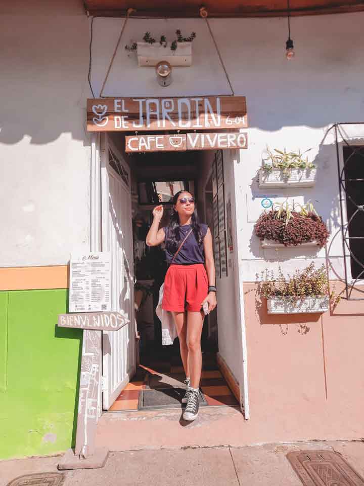 jardin-de-jardin A woman stands at the entrance of “El Café de Jardín,” a cozy café and plant shop in Jardín, Colombia, under a rustic wooden sign and surrounded by wall-mounted flower boxes.