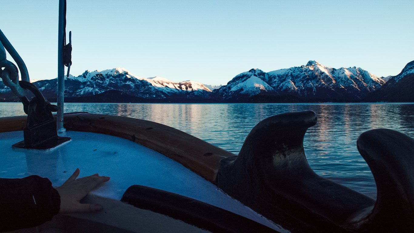 lake-nahuel-huapi-bariloche View from a boat on Lake Nahuel Huapi with snow-capped mountains glowing in the golden hour light and calm waters reflecting the scenic landscape.