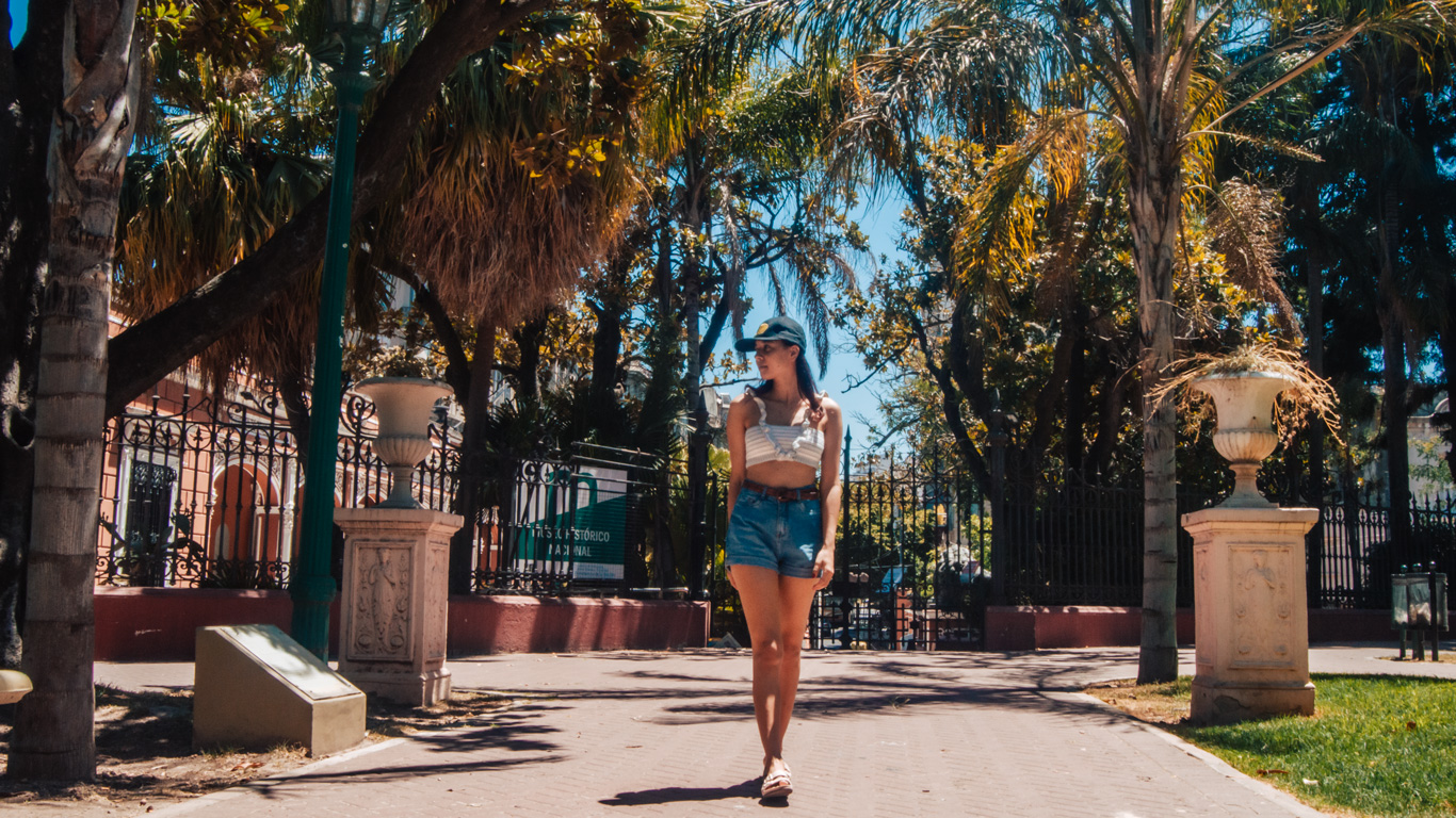 lezama-park-buenos-aires A woman in a crop top and denim shorts walks along a sun-dappled path lined with palm trees and classical urns in the tranquil Lezama Park in Buenos Aires.