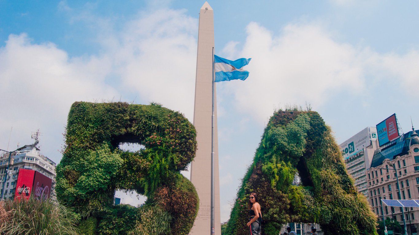 obelisco-de-buenos-aires-1 A woman poses in front of giant green letters "BA" made of plants, with the iconic Obelisco and Argentine flag behind her.