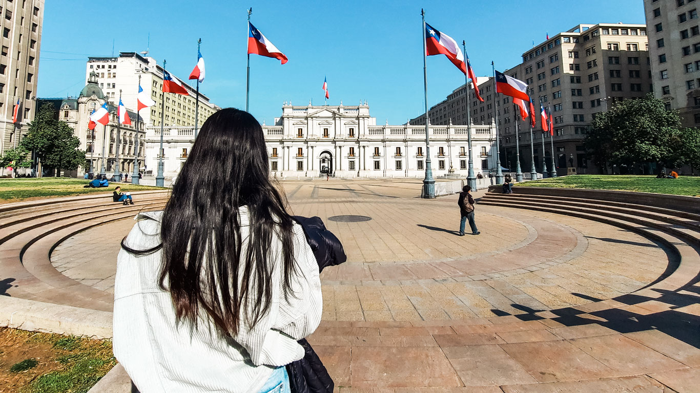 palacio-de-la-moneda-santiago A tourist looks toward Palacio de La Moneda, the presidential palace in Santiago, Chile, with Chilean flags waving under a clear blue sky.
