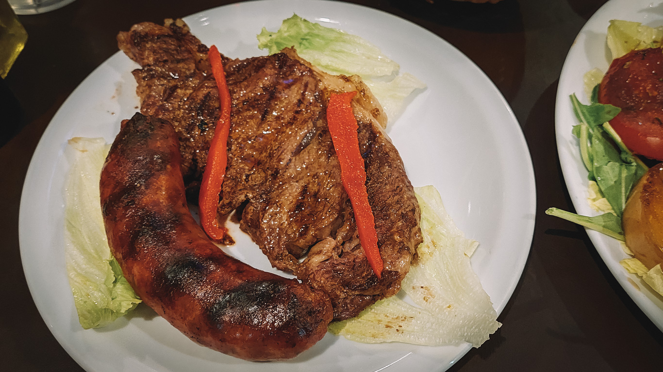parrilla-argentina A plate featuring a grilled ribeye steak with two strips of red bell pepper on top, served alongside a charred chorizo sausage and lettuce leaves.