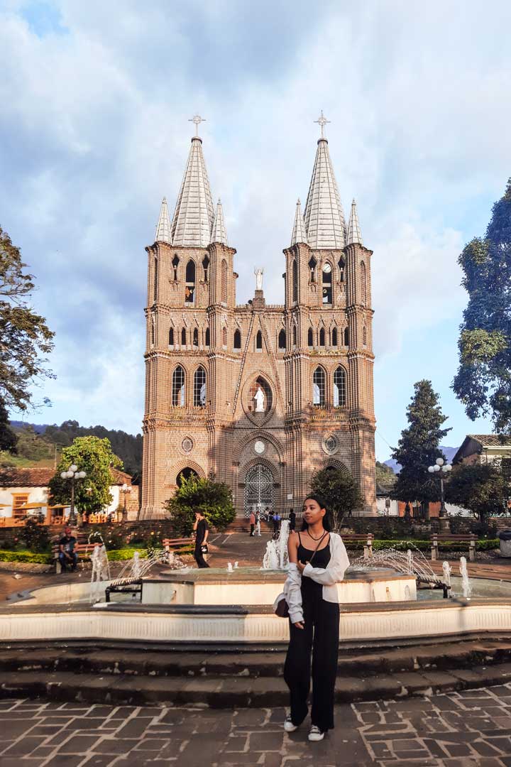 square-in-jardin A young woman stands in front of a large fountain in the central square of Jardín, Colombia, with the grand twin-spired Basilica of the Immaculate Conception rising in the background under a partly cloudy sky.