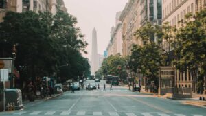 A view down Avenida 9 de Julio in Buenos Aires with the Obelisk centered in the distance, framed by trees and classic architecture as people cross the street.