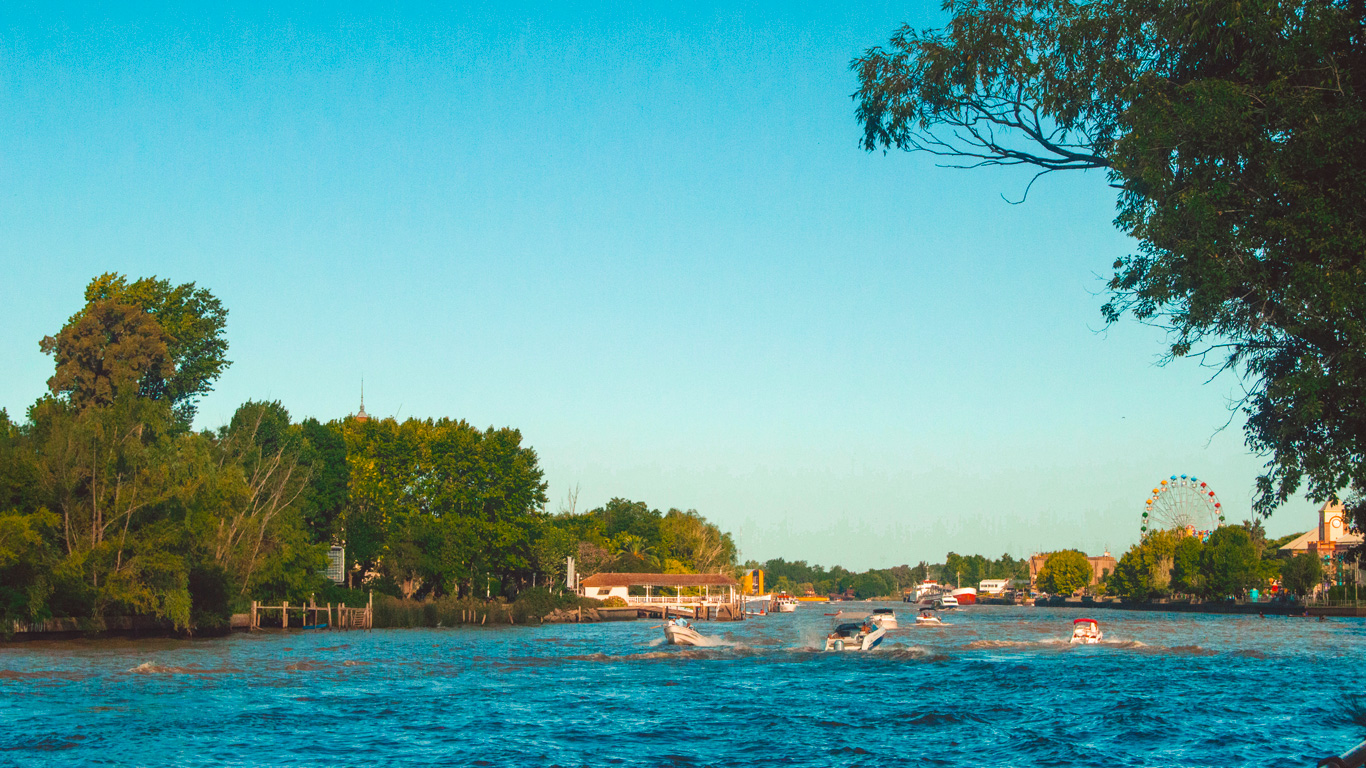 tigre-buenos-aires-1 A wide river channel with several speedboats cruising through it, surrounded by lush green trees and a distant Ferris wheel on the right, under a clear blue sky.