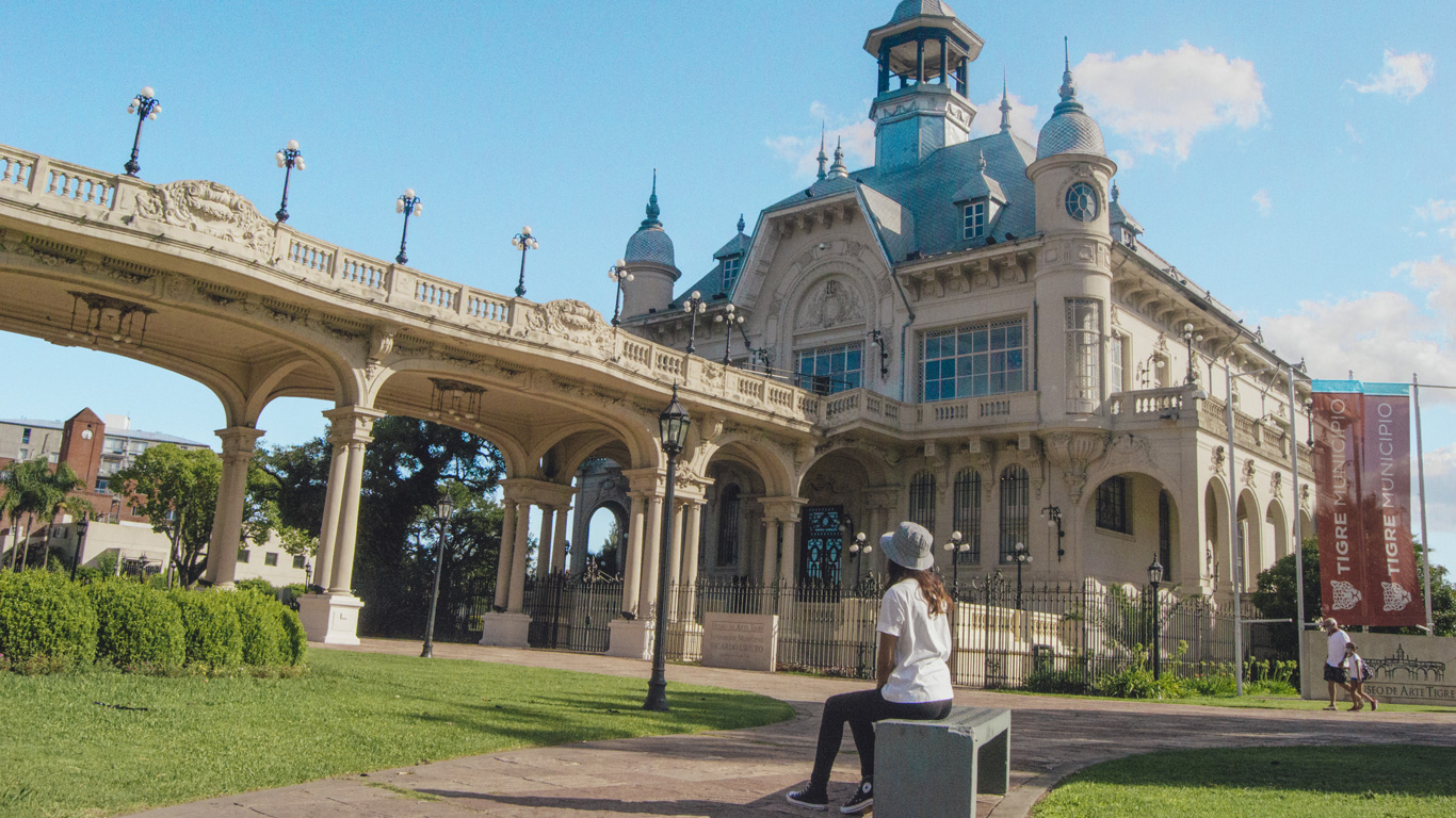 tigre-buenos-aires A person wearing a white hat and t-shirt sits on a bench admiring the ornate European-style architecture of the Museo de Arte Tigre, under a bright blue sky.