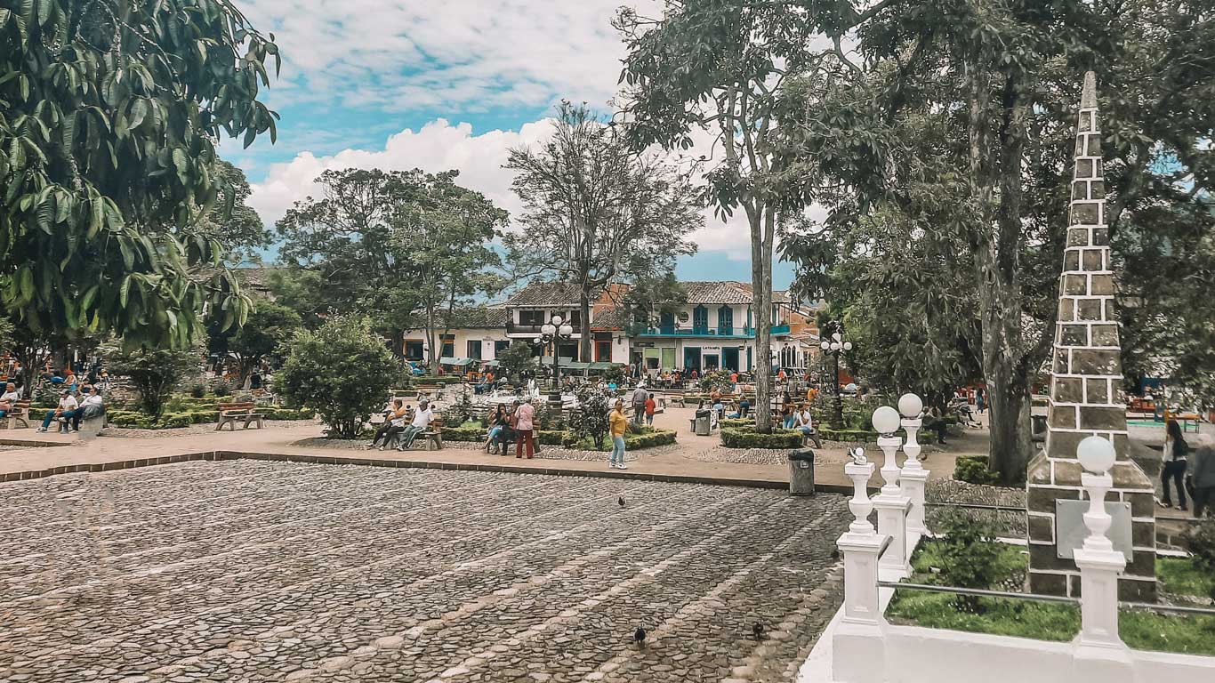 transportation-in-colombian A bustling town square in Jardín Antioquia, filled with people relaxing on benches and strolling through tree-lined paths surrounded by colonial buildings. The foreground features a patterned cobblestone area and a white stone monument with spherical lamps, adding charm to the vibrant public space.
