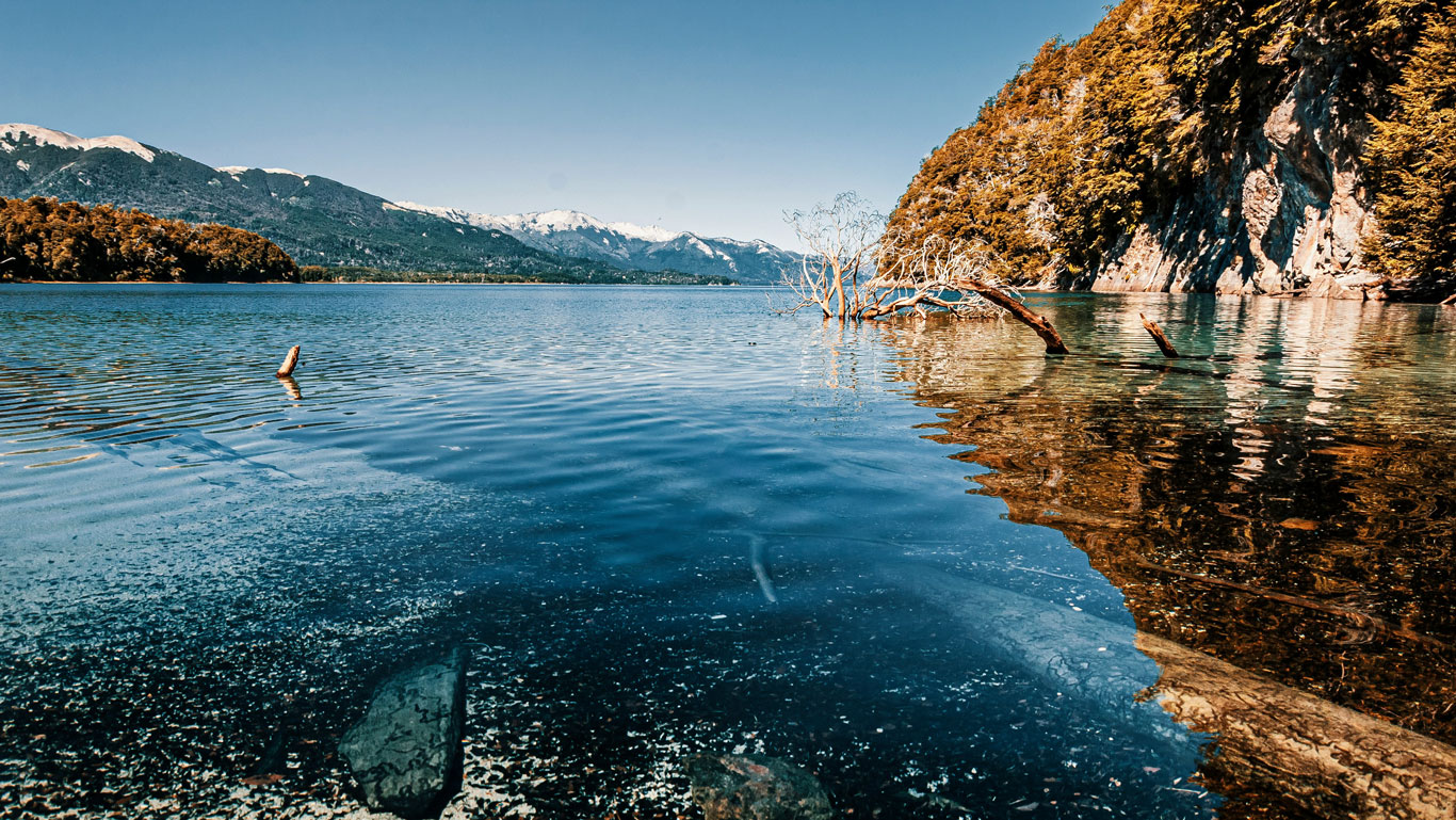 villa-la-angostura Crystal-clear waters reflecting forested cliffs and snow-capped mountains in Villa La Angostura, a must-see stop near the snow in Bariloche.