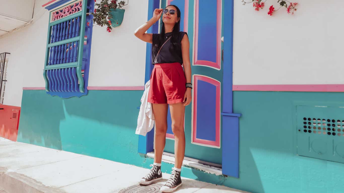 A woman in red shorts and black high-top sneakers stands confidently in front of a colorfully painted door and window in Jardín, Colombia, with vibrant teal, pink, and purple details popping in the bright sunlight.