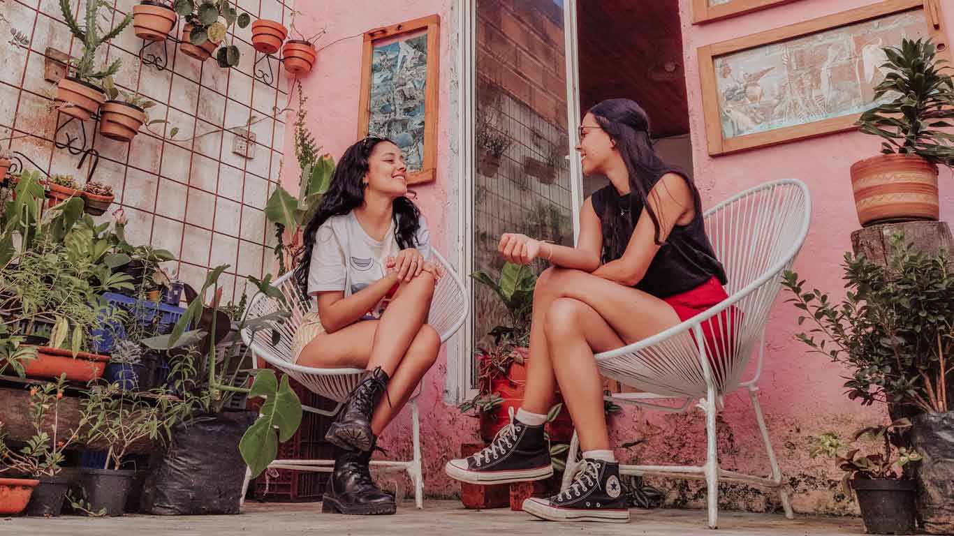 what-to-do-in-jardin Two women sit across from each other in white patio chairs, laughing and chatting in a cozy courtyard café in Jardín, Colombia, surrounded by potted plants and rustic decor.