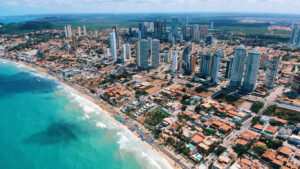 Panoramic aerial photo of Natal’s coastline showing dense clusters of tall buildings inland and a lively beachfront with turquoise waters and a long line of umbrellas and beach huts.