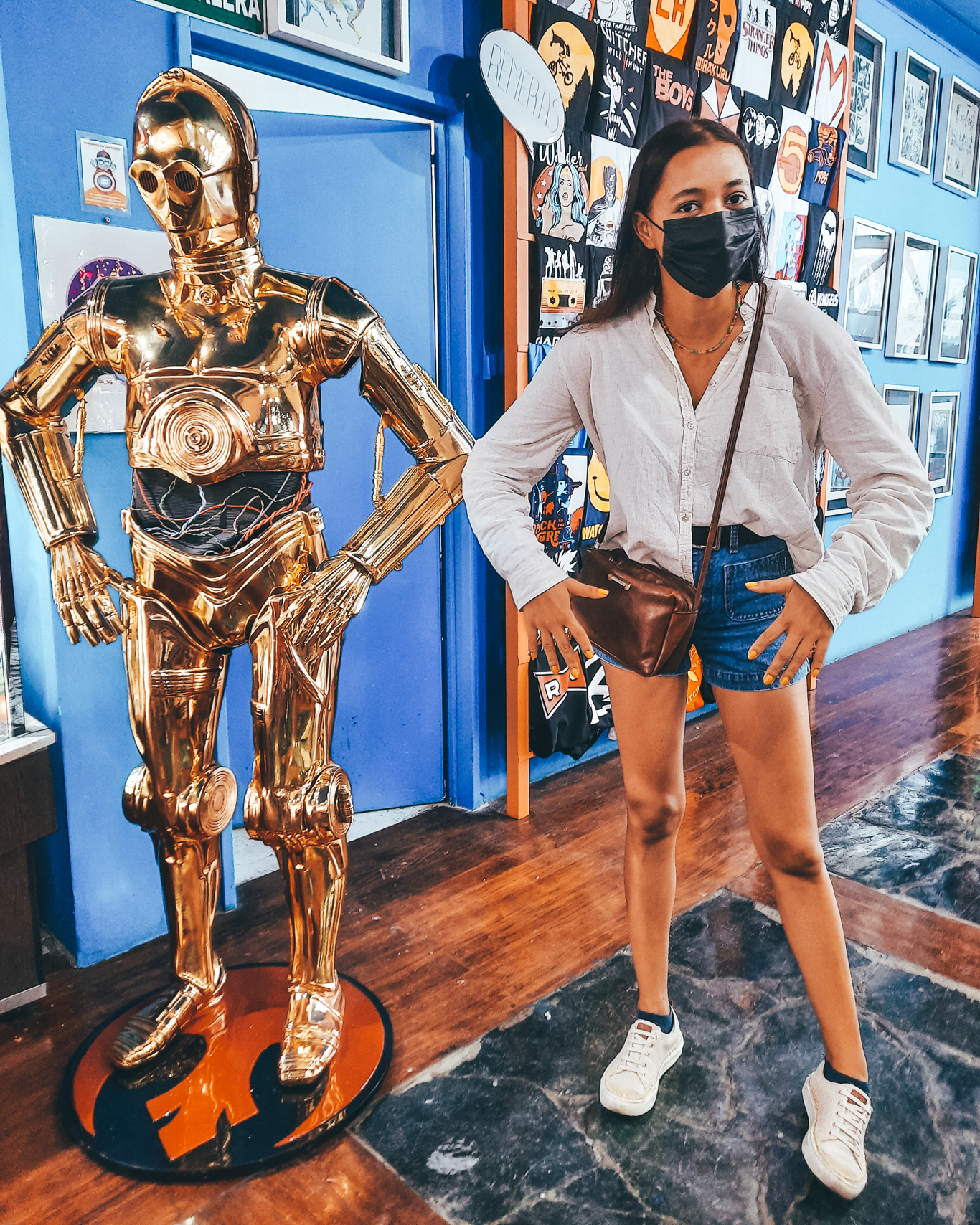 La-Revisteria-Buenos-Aires-1 A woman wearing a mask poses humorously beside a golden C-3PO statue inside La Revistería comic shop in Buenos Aires. Behind them are graphic posters and pop culture-themed merchandise on brightly colored walls.