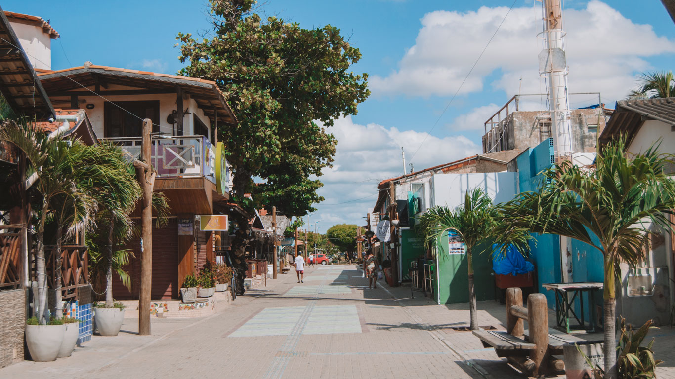 canoa-quebrada-brazil-2 Charming and quiet street with rustic-style shops and inns, lined with palm trees and trees under a blue sky with clouds, in the downtown area, the best place to stay in Canoa Quebrada. A few people walk in the background in a relaxed atmosphere.