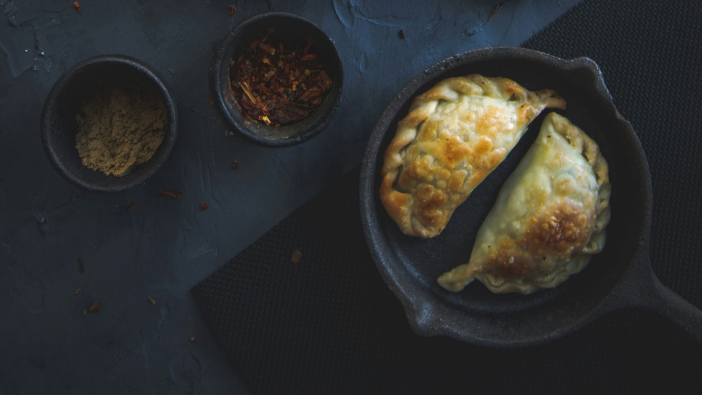 cuisine-ushuaia Two golden, flaky empanadas sit in a cast iron skillet on a dark slate surface, flanked by small bowls of ground spices and crushed red pepper. The moody lighting highlights the rustic and hearty appeal of Argentine cuisine.