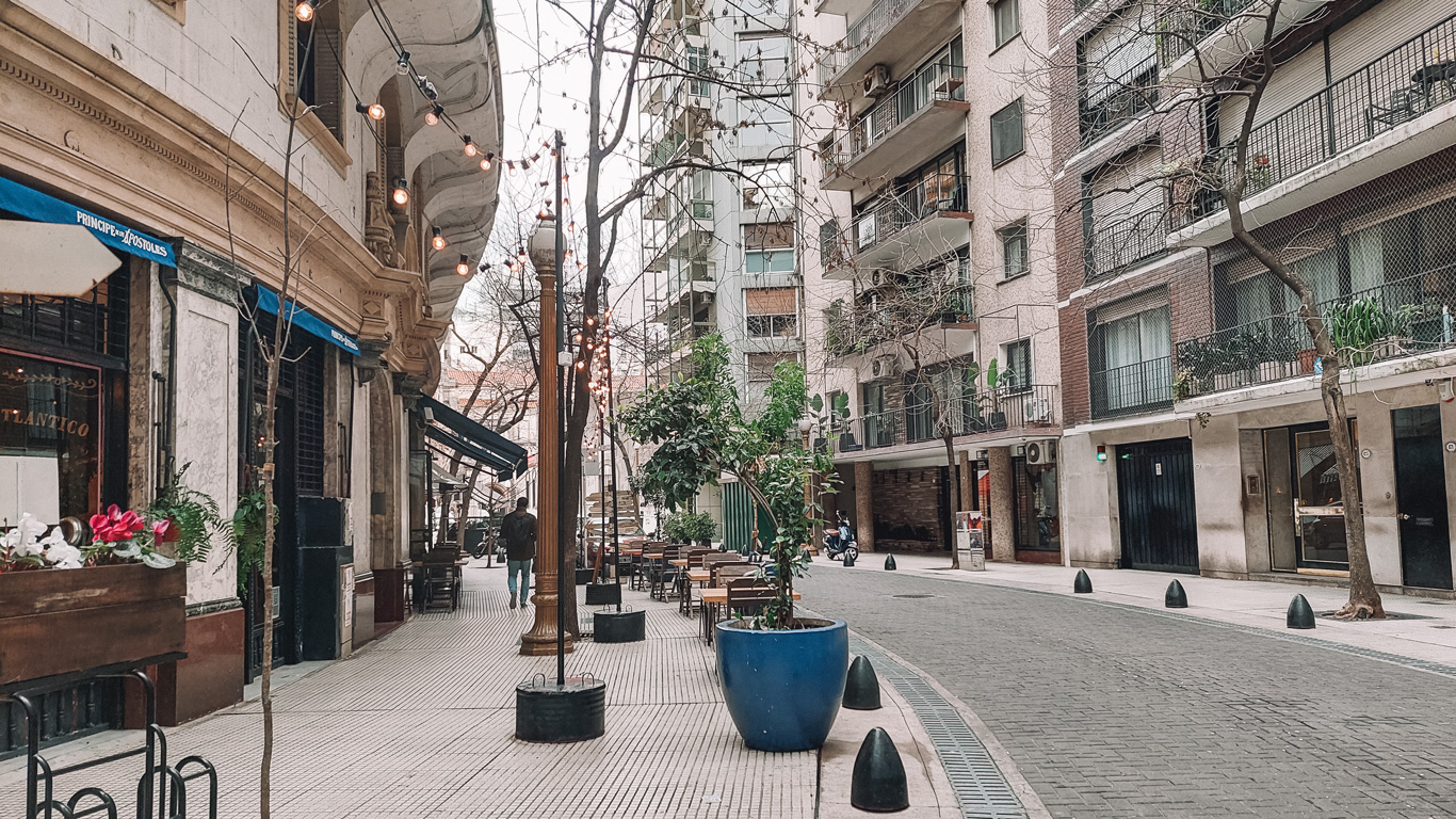 floreria-atlantico-buenos-aires A stylish café scene in Buenos Aires featuring outdoor seating with string lights, potted plants, and an old-world building with blue awnings labeled "Principe de los Apostoles." A quiet, cozy street sets a relaxed ambiance.
