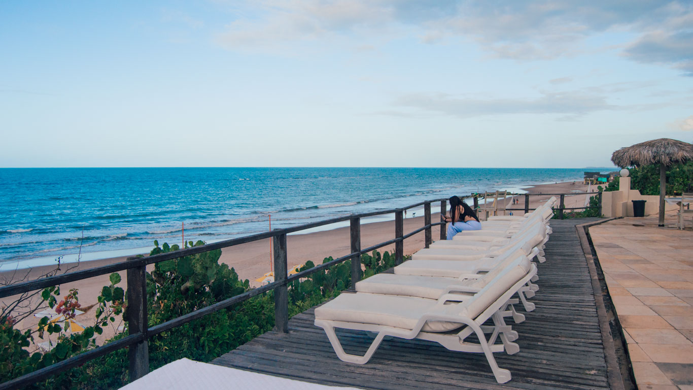 hotels-in-canoa-quebrada Wooden deck with white lounge chairs facing the beach at Pousada Vila Canoa, where a woman sits looking at the blue sea under a clear sky. This is an excellent choice of where to stay in Canoa Quebrada.
