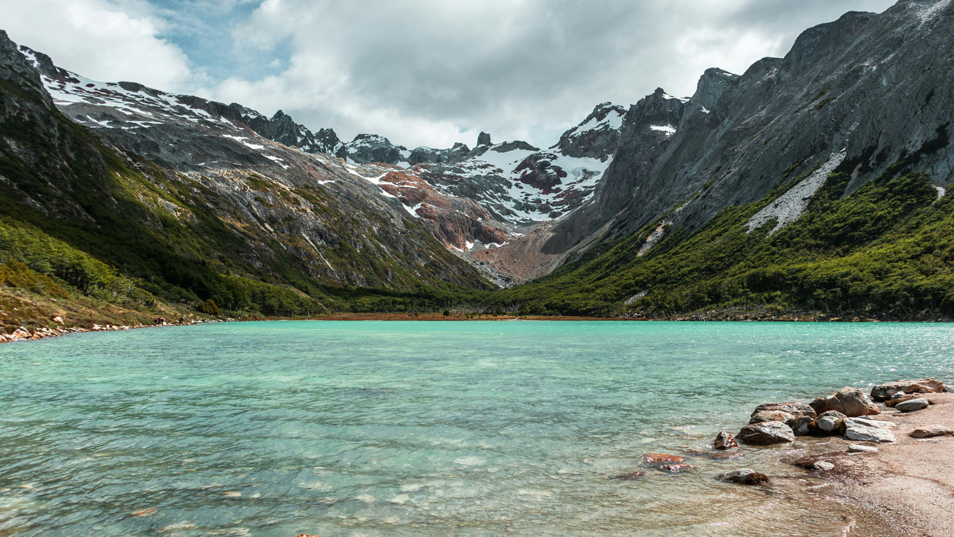 laguna-esmeralda-ushuaia A turquoise glacial lake known as Laguna Esmeralda sits in the middle of a dramatic valley, surrounded by rocky slopes, green forest, and snow-capped peaks. The cloudy sky enhances the contrast of colors in this stunning natural landmark near Ushuaia.