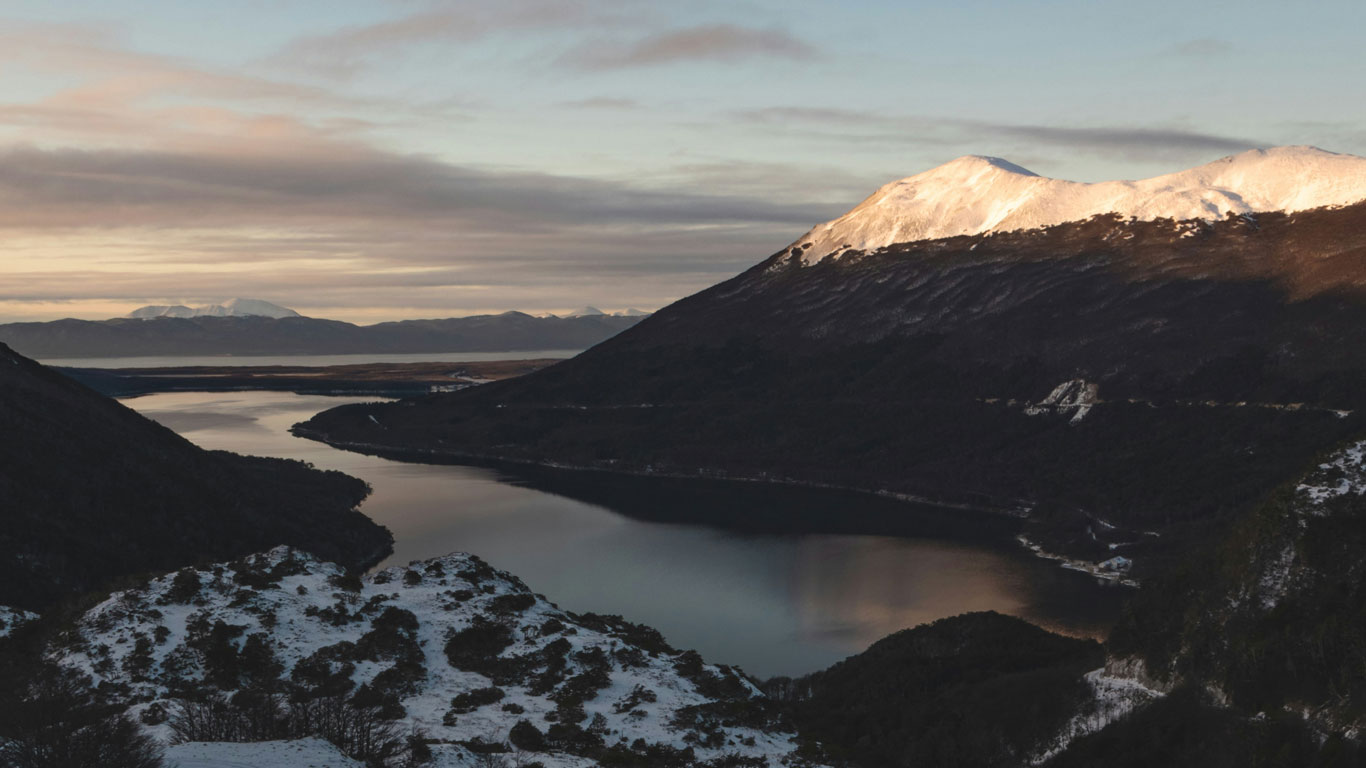 lake-escondido-ushuaia A serene alpine lake winds through snow-dusted hills and dark forested mountains, with golden sunlight illuminating the distant peaks. The tranquil scene captures the dramatic, untouched landscape near Lake Escondido in Ushuaia.