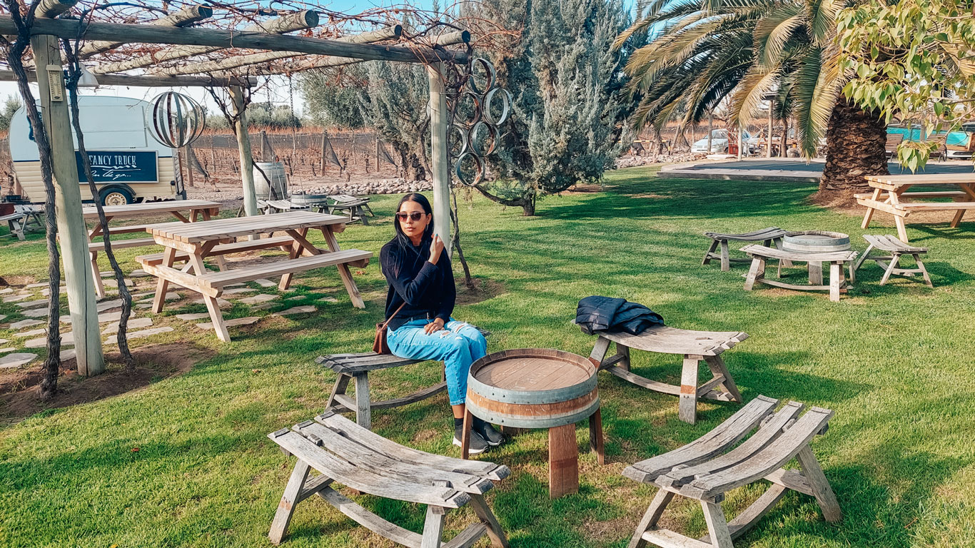 Woman sitting in a well-maintained garden at the Tempus Alba winery in Mendoza, surrounded by rustic wooden benches and tables, with a trailer in the background.