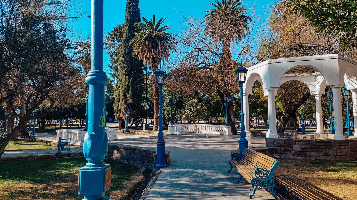 Independence Square in Mendoza in October, tree-lined with wooden benches, blue lampposts, and a white gazebo, under a clear blue sky.