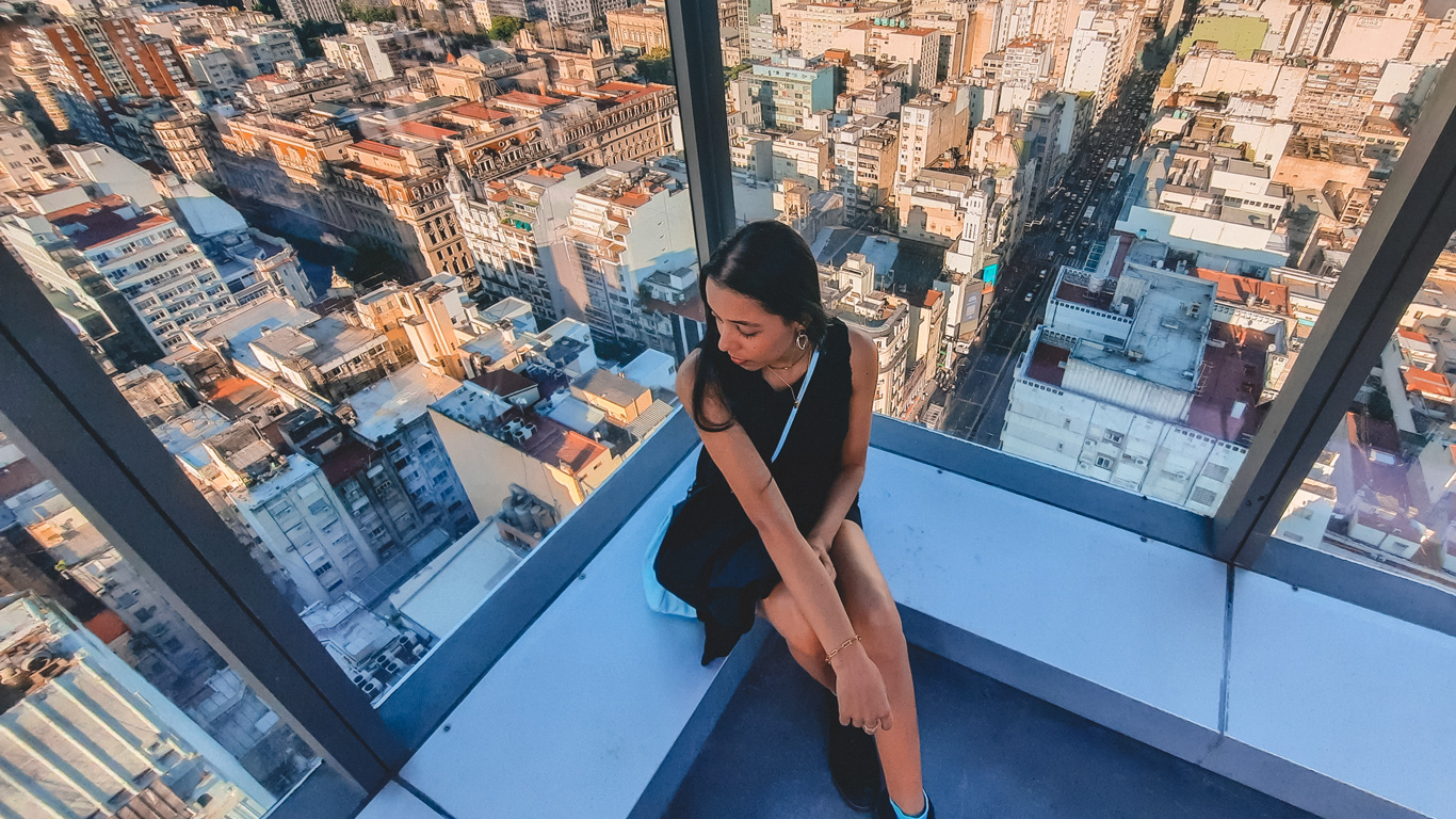 olympo-sky-bar-buenos-aires A woman in a black dress sits near the glass wall of a high-rise bar, overlooking a panoramic cityscape of Buenos Aires with densely packed buildings stretching into the distance. The view captures the urban texture and vibrant sprawl of the Argentine capital.