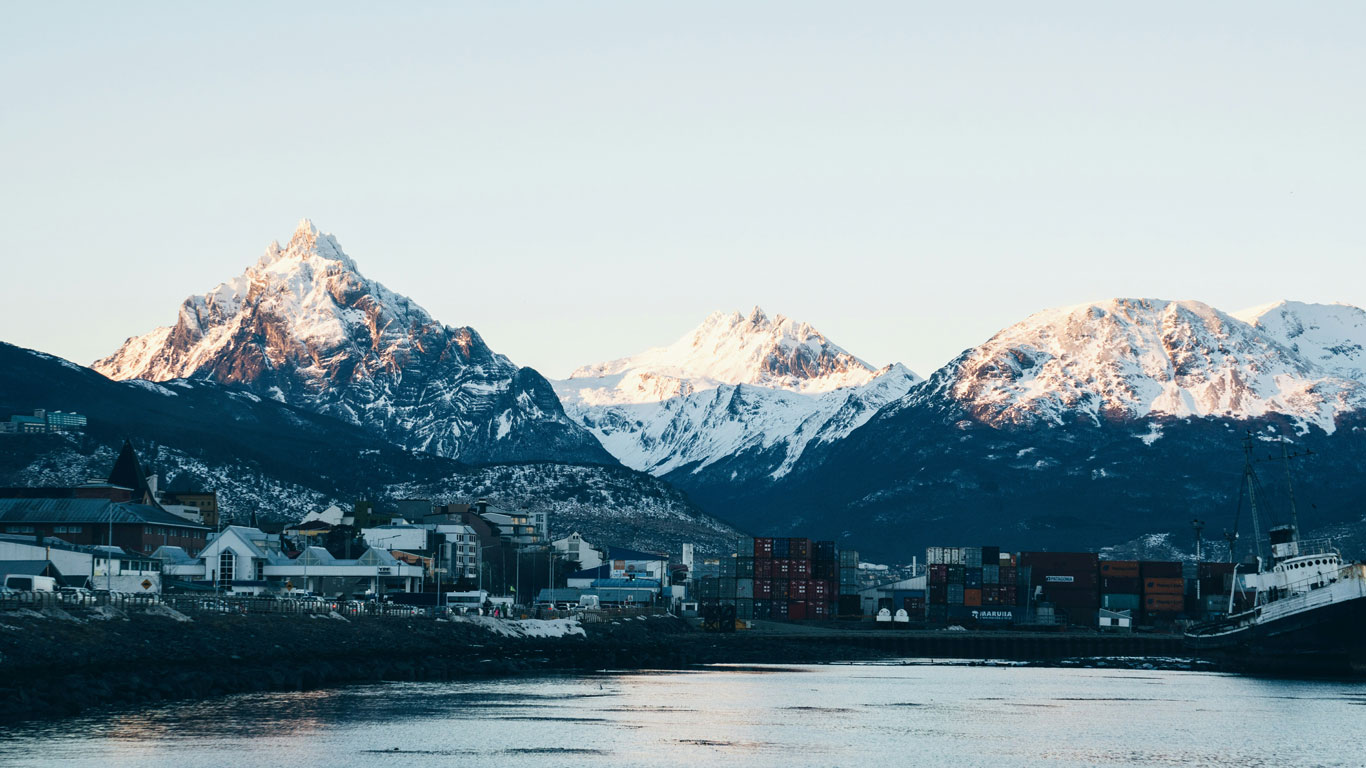 snow-in-ushuaia-argentina Ushuaia’s cityscape sits along a calm waterfront, with snow-covered mountains rising dramatically in the background under a soft, clear sky. Shipping containers, buildings, and a docked ship appear below jagged, sunlit peaks.
