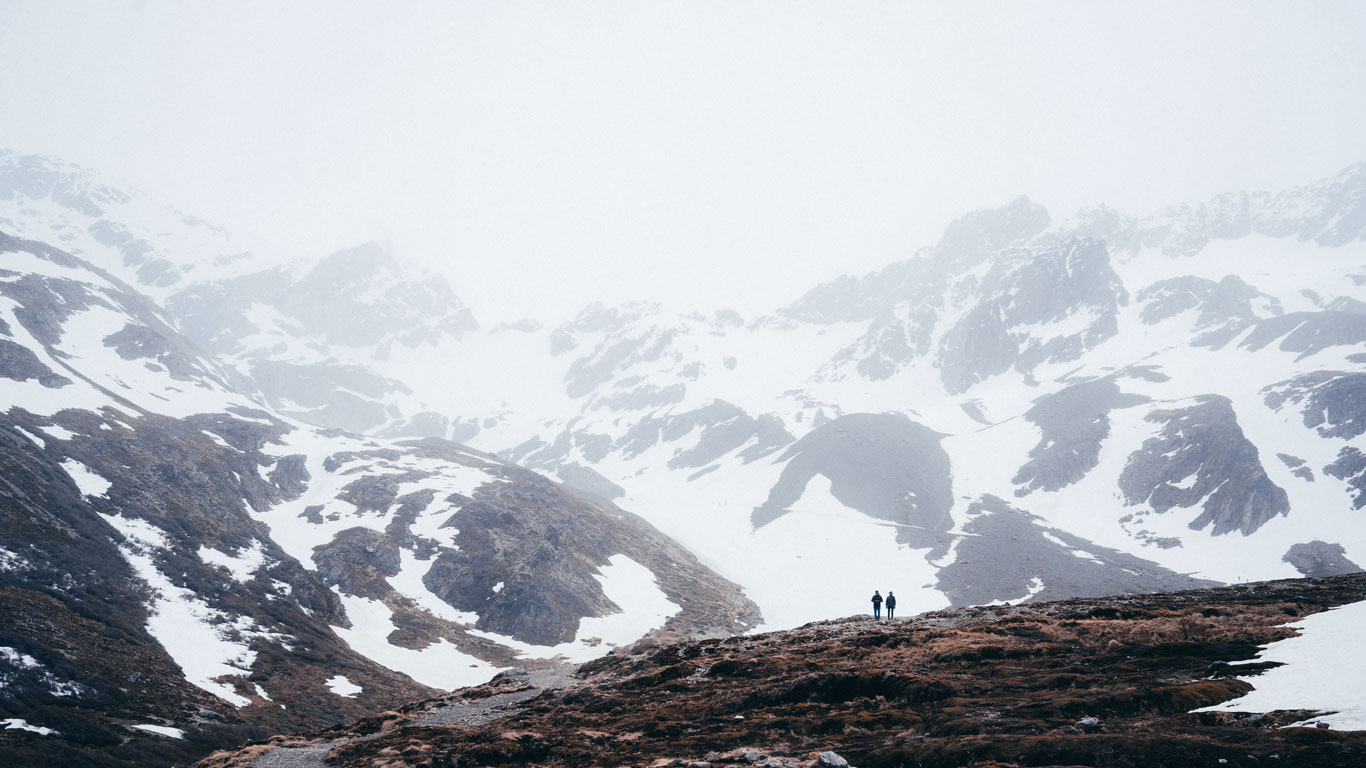 Two hikers stand on a rugged, snow-dusted mountain trail, dwarfed by towering, mist-covered peaks and scattered patches of snow. The foggy atmosphere and stark contrast between rock and snow create a moody, dramatic landscape typical of Ushuaia’s remote wilderness.