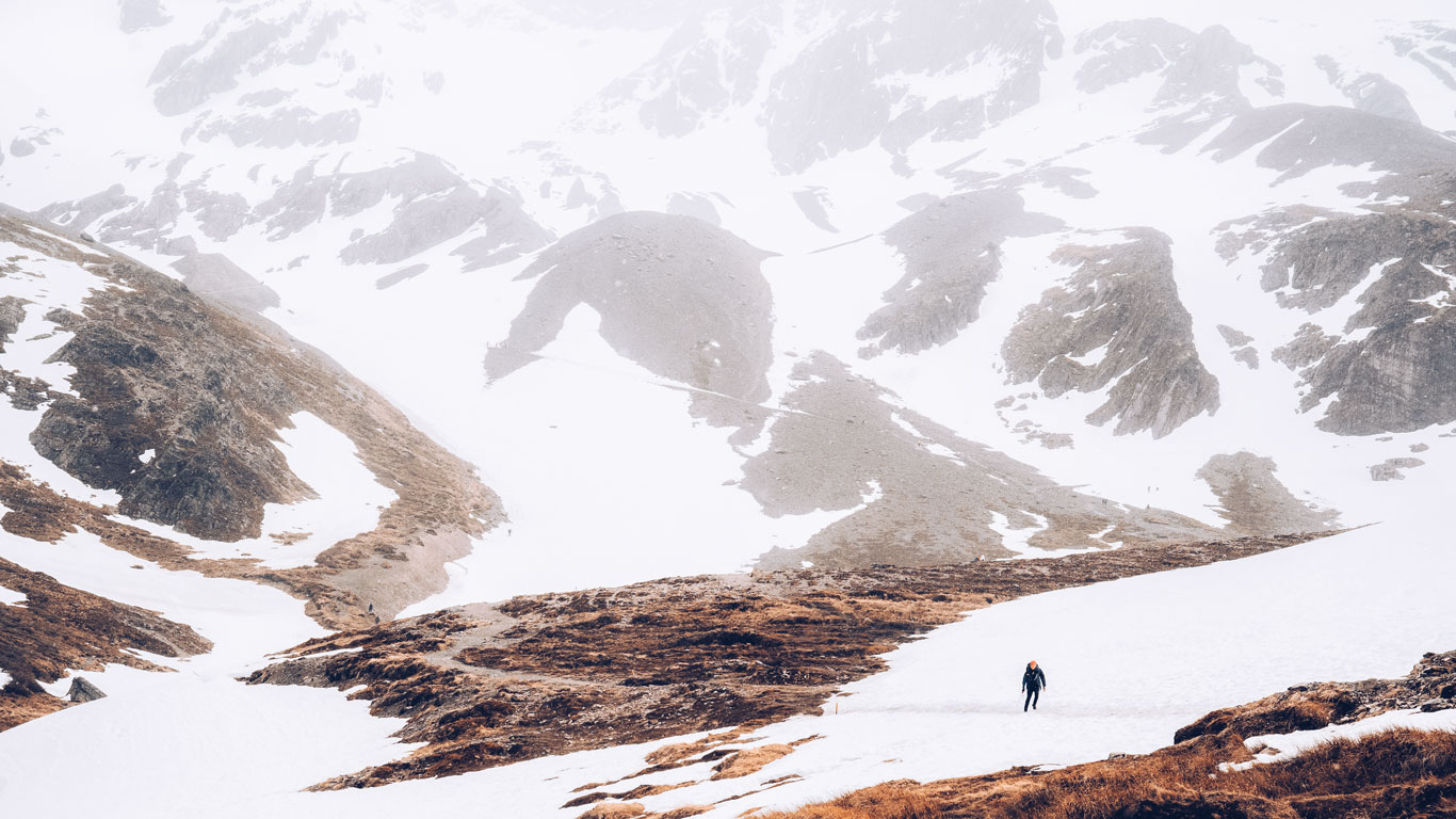 snow-ushuaia A lone hiker in dark clothing walks up a snowy incline, surrounded by vast, snow-covered peaks and steep, barren slopes. The expansive view emphasizes the remote and rugged terrain of Ushuaia’s highlands.