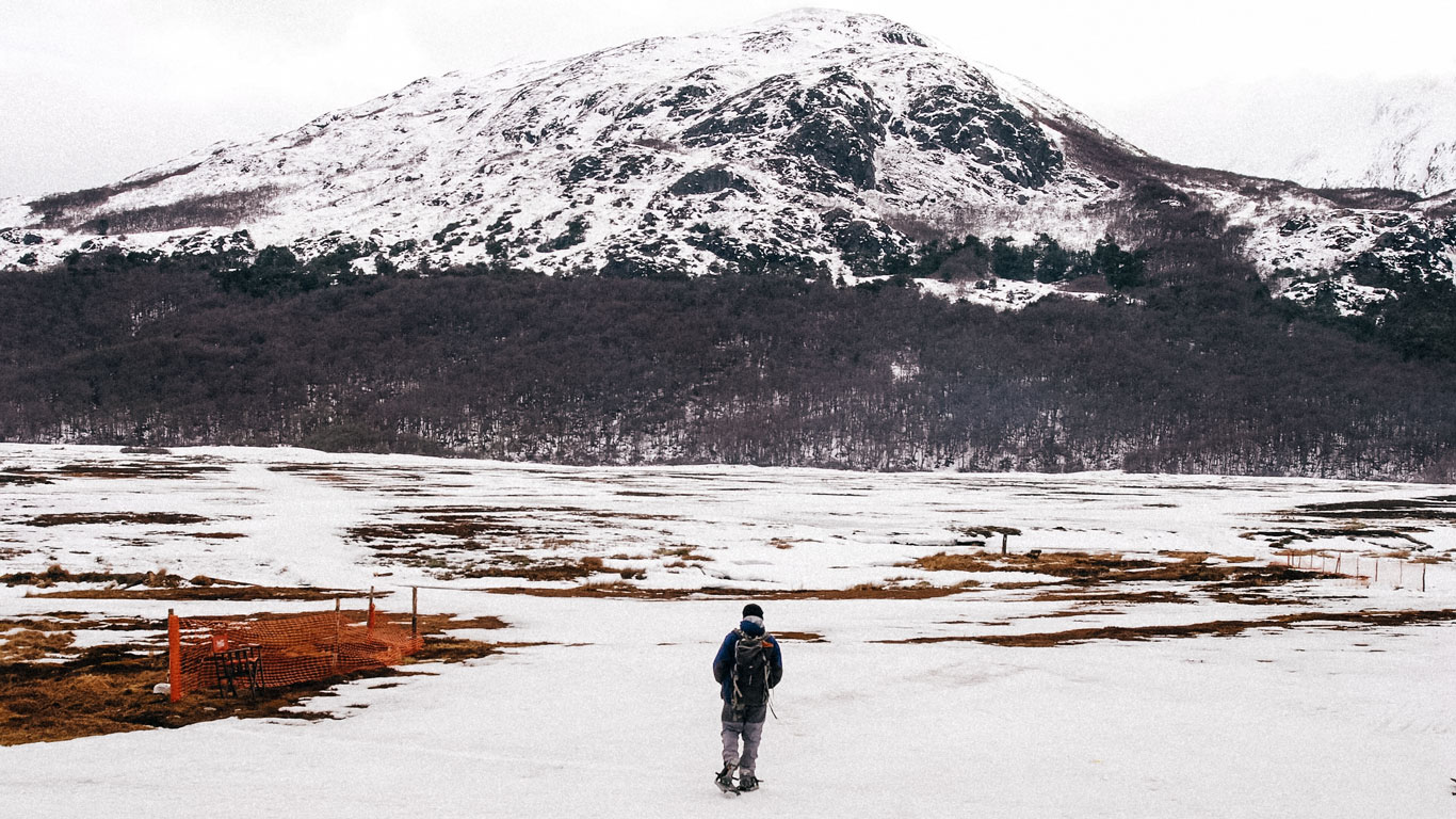 snowshoe-hiking-ushuaia A person wearing snowshoes and a backpack walks alone through a snow-covered valley, facing a tree-lined mountain in the distance. The open, wintry terrain conveys the solitude and adventure of snowshoe hiking in Ushuaia, Argentina.
