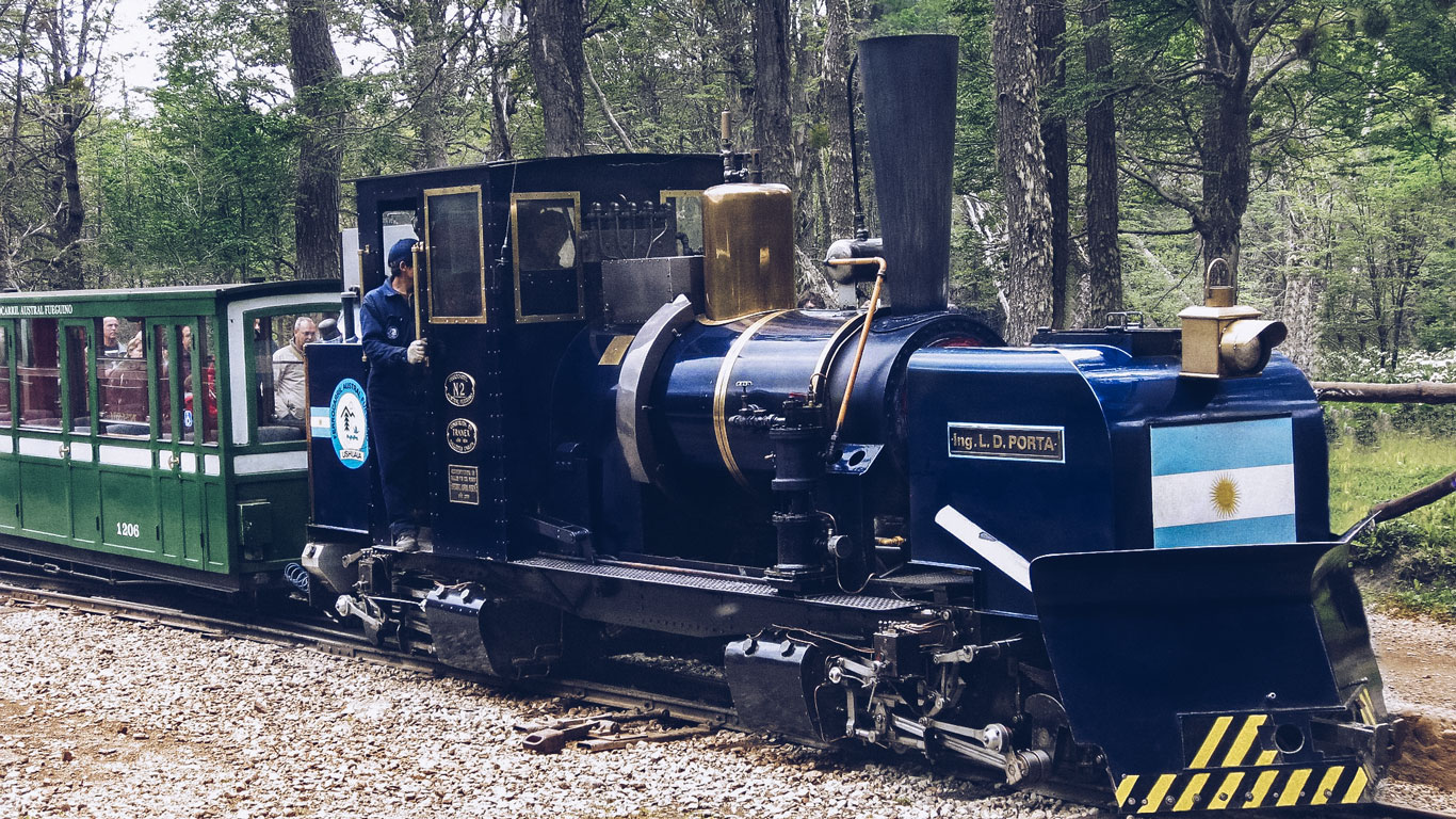 the-end-of-the-world-train-ushuaia A deep blue steam locomotive with gold accents and an Argentine flag pulls green passenger cars through the forest. Known as the End of the World Train, it carries tourists through southern Patagonia’s rugged wilderness near Ushuaia.