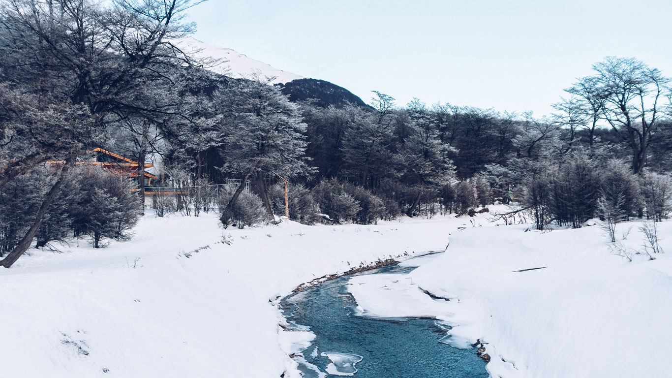 tierra-del-fuego-national-park-ushuaia A narrow, partially frozen stream winds through a snow-covered forest at the base of the mountains in Tierra del Fuego National Park. Snow-laden trees and a wooden structure partially hidden in the background capture the park’s serene, wintry beauty.