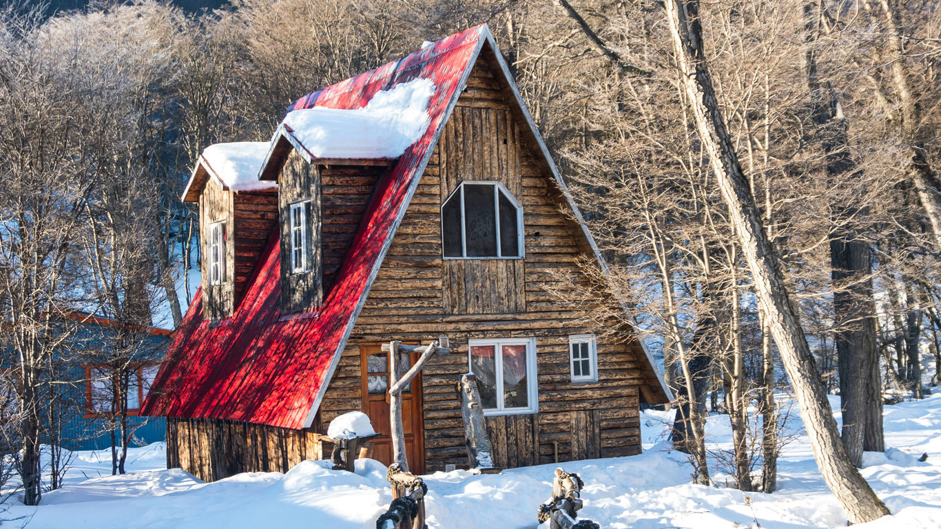 ushuaia-argentina A rustic wooden A-frame cabin with a steep red metal roof sits surrounded by bare trees and deep snow. Sunlight highlights the charm and warmth of this cozy forest retreat in Ushuaia’s winter landscape.