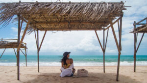 Woman sitting on the sand under a rustic wooden beach hut with a thatched roof, looking at the blue sea of Canoa Quebrada. Next to her, a beach bag and flip-flops, in a peaceful seaside atmosphere.