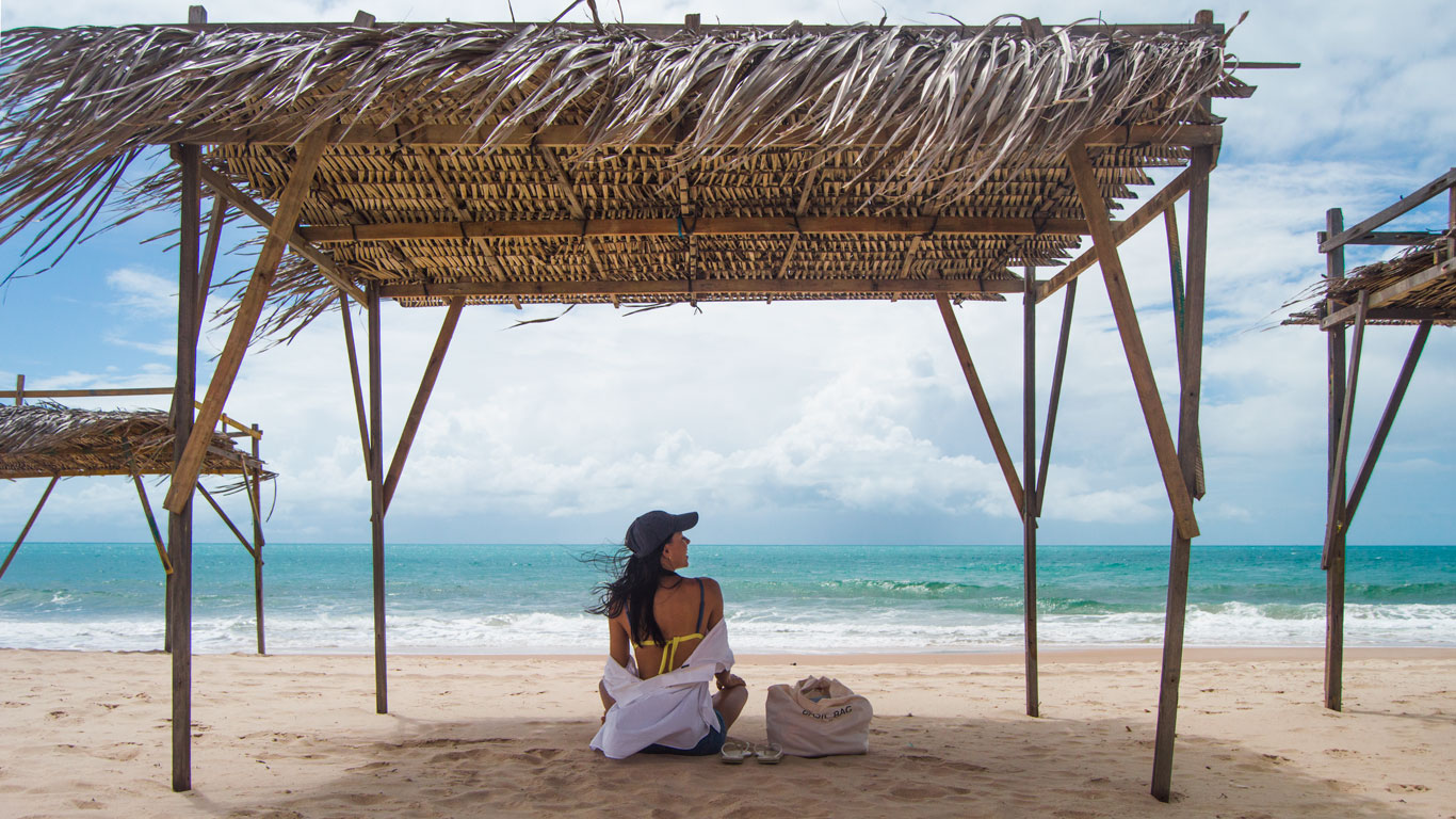 Woman sitting on the sand under a rustic wooden beach hut with a thatched roof, looking at the blue sea of Canoa Quebrada. Next to her, a beach bag and flip-flops, in a peaceful seaside atmosphere.