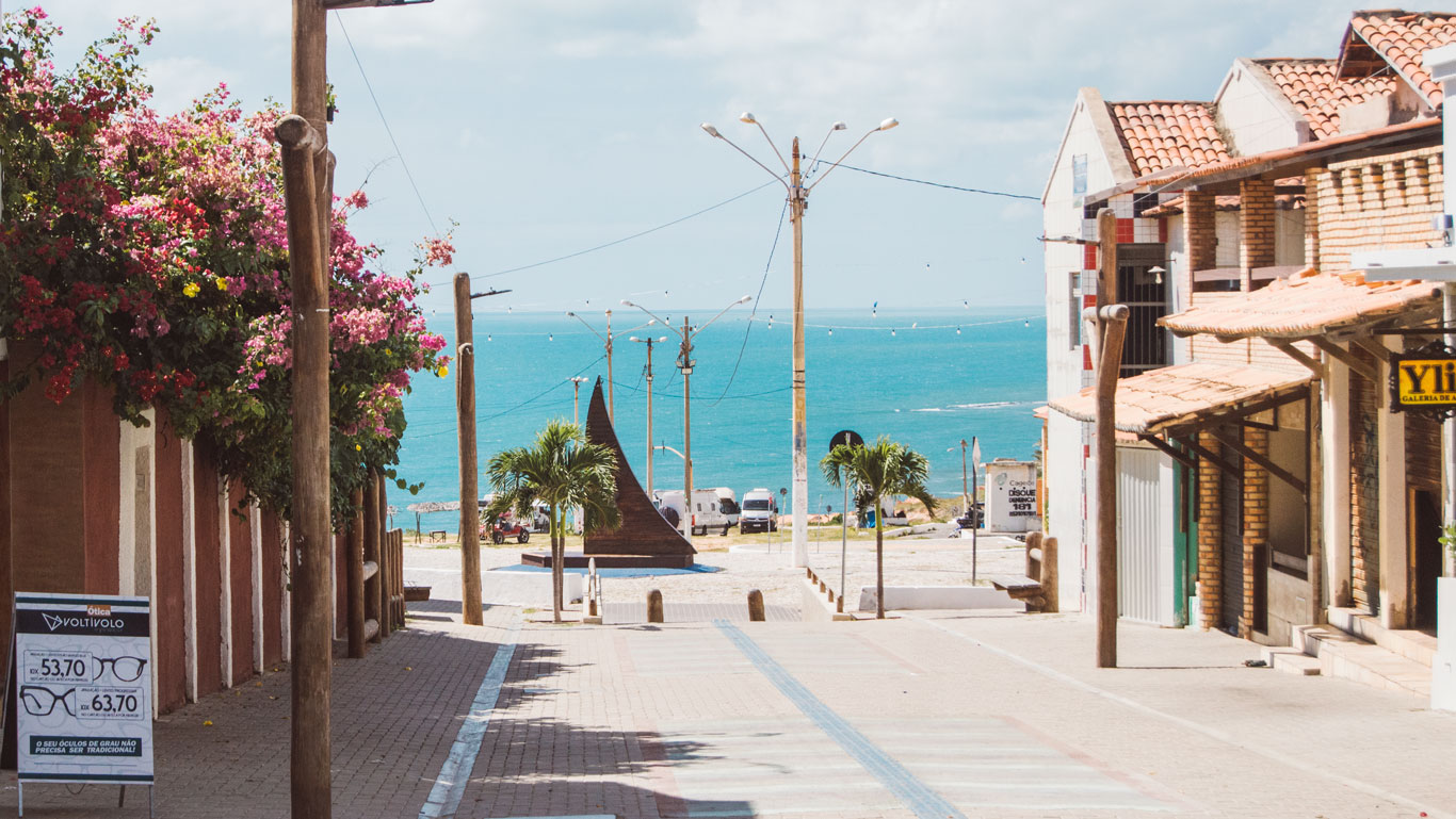 where-to-stay-in-canoa-quebrada View of Broadway Street in downtown Canoa Quebrada, sloping down toward the blue sea, with lampposts, palm trees, and some brick buildings with commercial signs. A sailboat sculpture appears in the background.