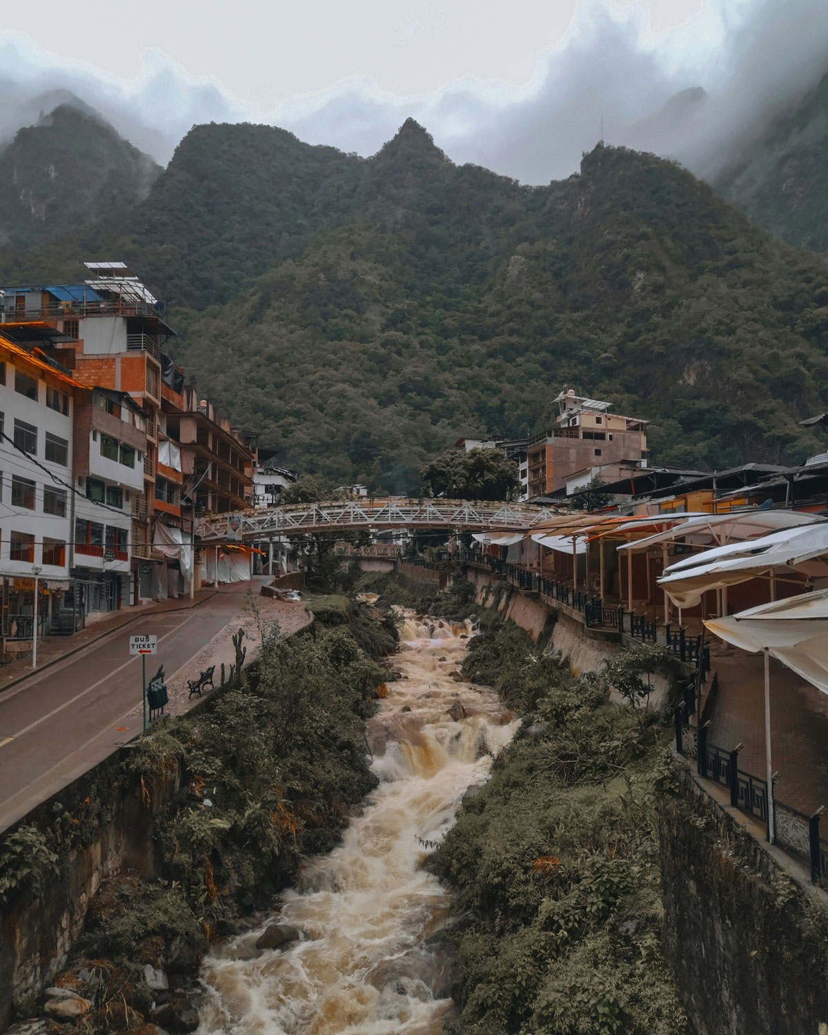 Aguas Calientes, the gateway town to Machu Picchu, with rustic buildings lining a rushing river and misty green mountains in the background.