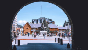 Bariloche’s central square seen through a stone arch, with people walking around in winter clothes. In the background, charming wooden and stone buildings frame the scene, while a horse sculpture stands prominently at the center. The sky is clear and blue, revealing a cold yet sunny day.