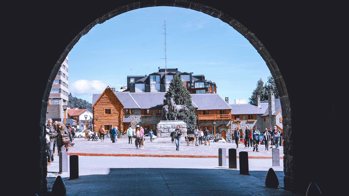 Bariloche’s central square seen through a stone arch, with people walking around in winter clothes. In the background, charming wooden and stone buildings frame the scene, while a horse sculpture stands prominently at the center. The sky is clear and blue, revealing a cold yet sunny day.