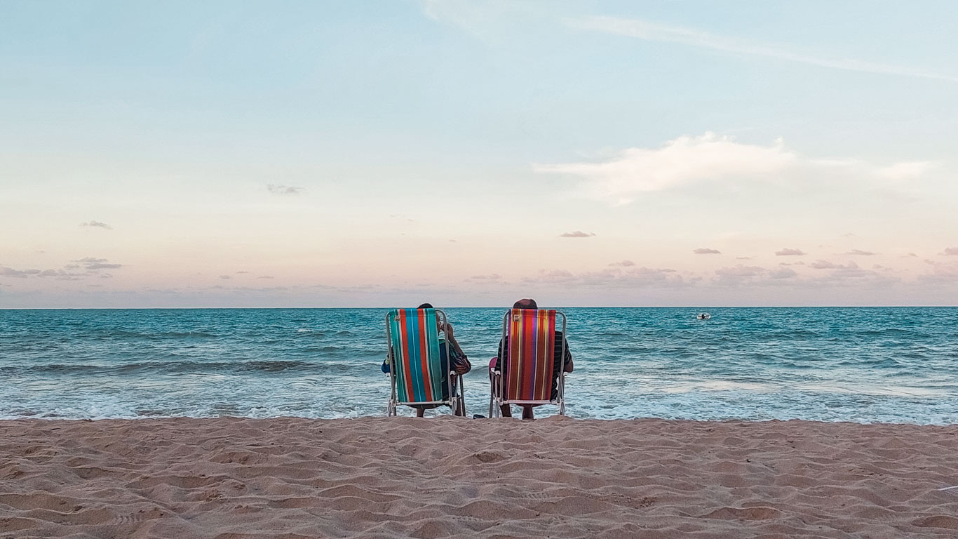 Two people sit in colorful beach chairs facing the calm sea at sunset on Bessa Beach, under a sky painted in soft pastel hues.