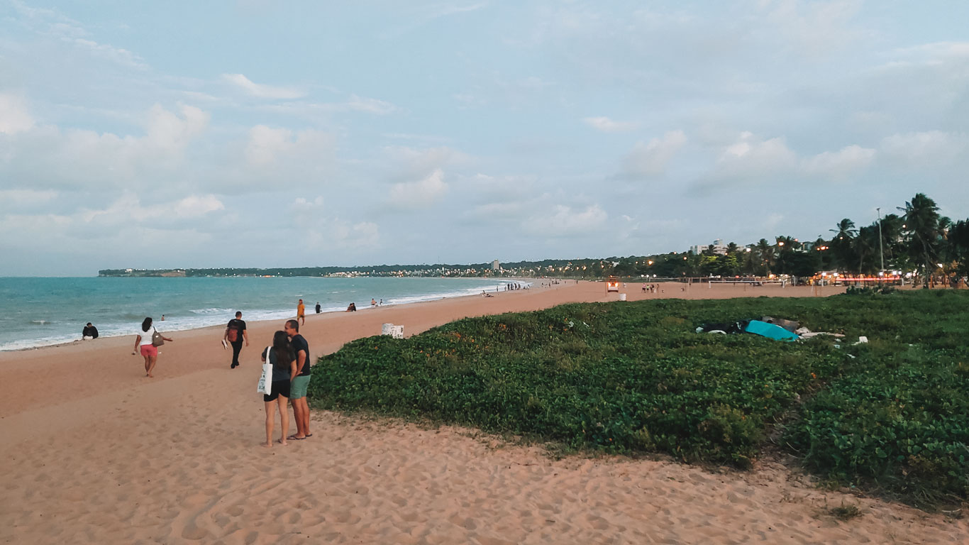 People walking and chatting along the sandy stretch of Cabo Branco Beach, with low coastal vegetation to the right and the cityscape of João Pessoa in the background.