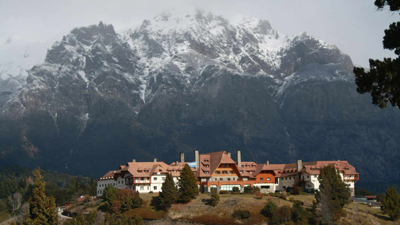 Panoramic view of a majestic snow-covered mountain with the grand Llao Llao Resort in Bariloche nestled among trees and lush greenery.