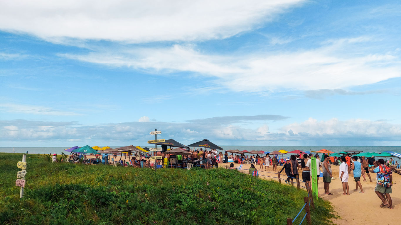 Bustling Jardim Oceania Beach, with colorful beach stalls and people milling about, next to a grassy area with signs pointing out directions.