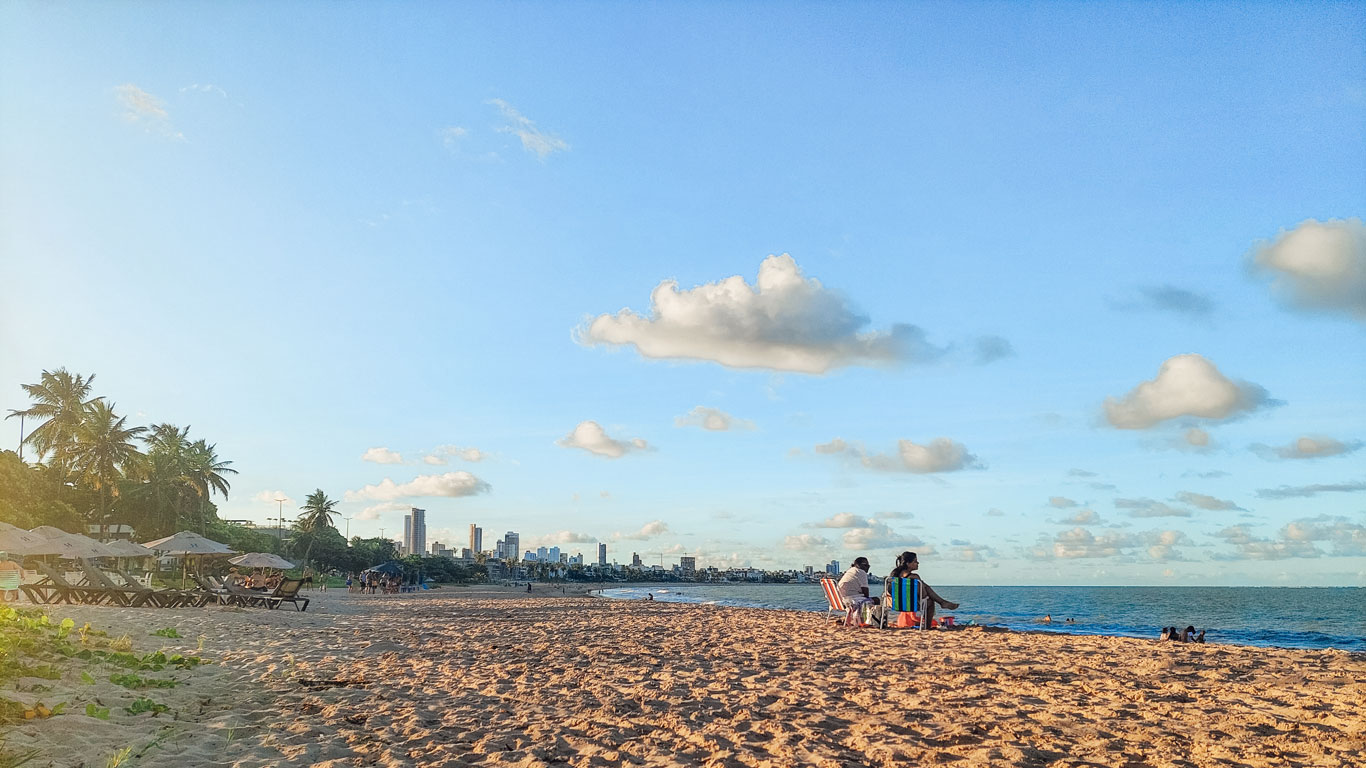 Manaíra Beach with palm trees and empty beach chairs on the left, while a few people unwind on the sand to the right. In the background, João Pessoa’s skyline rises beneath a golden-blue late afternoon sky.